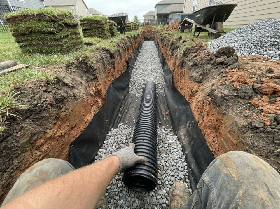 Open French drain trench in a lawn lined with filter fabric, with black perforated pipe on washed gravel and clay subsoil in the trench walls